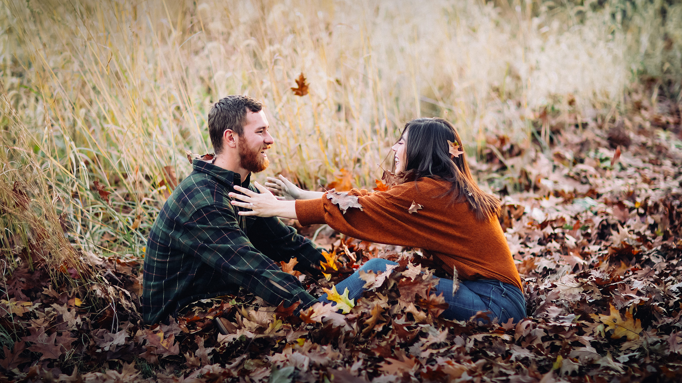 an engaged couple sitting in a pile of fall leaves