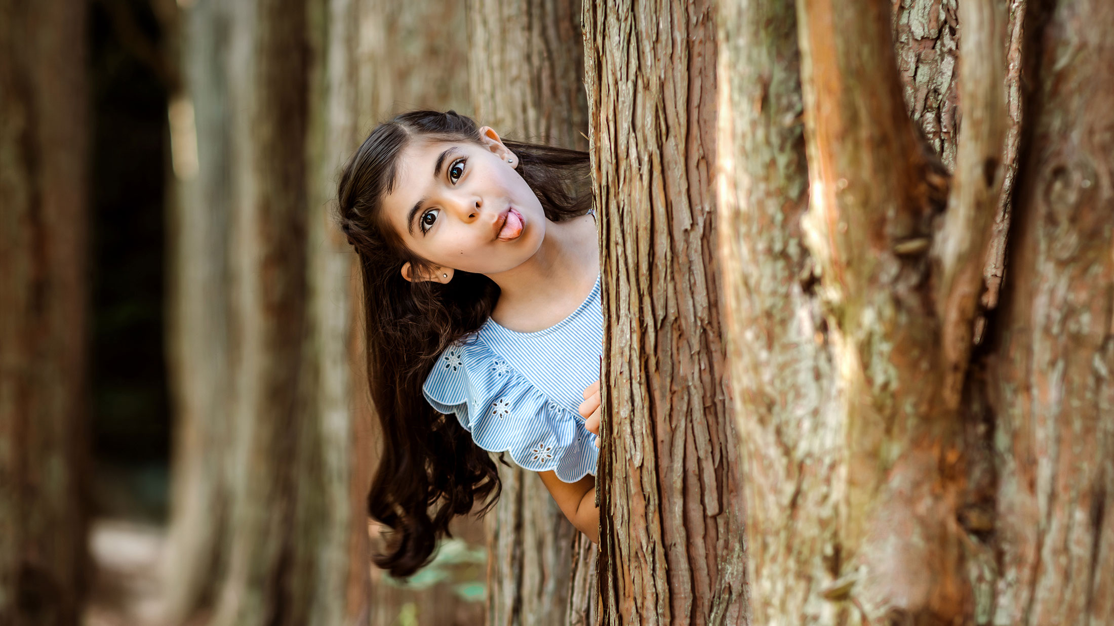 Little girl peeking from between trees and sticking out her tongue